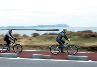 Two bicycle riders riding on a bicycle path with the sea in the background